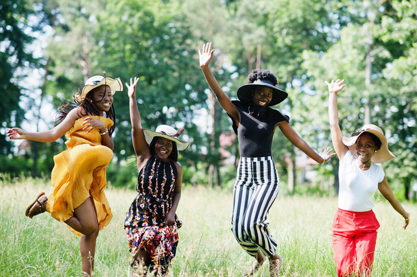 Group of four gorgeous african american womans wear summer hat jumping at green grass in park. - Φωτογραφία, εικόνα