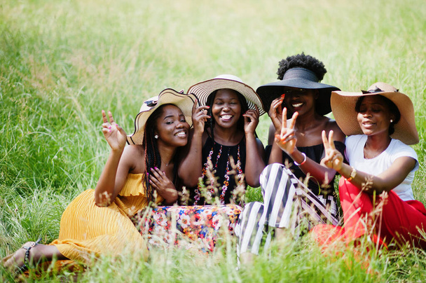 Dört muhteşem Afrikalı Amerikalı kadın grubu parkta yeşil çimlerde oturan yaz şapkası takıyor.. - Fotoğraf, Görsel
