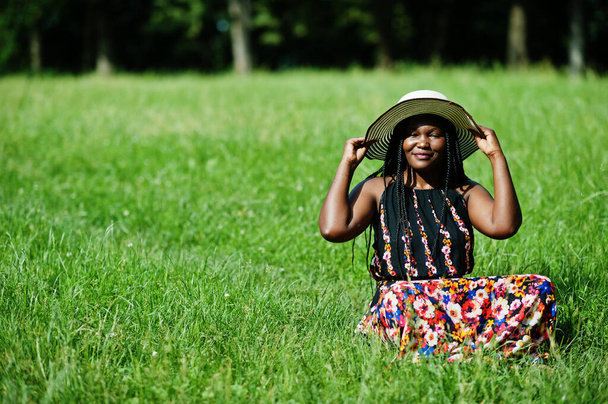Portrait of gorgeous african american woman 20s in summer hat sitting at green grass in park. - Foto, Imagem