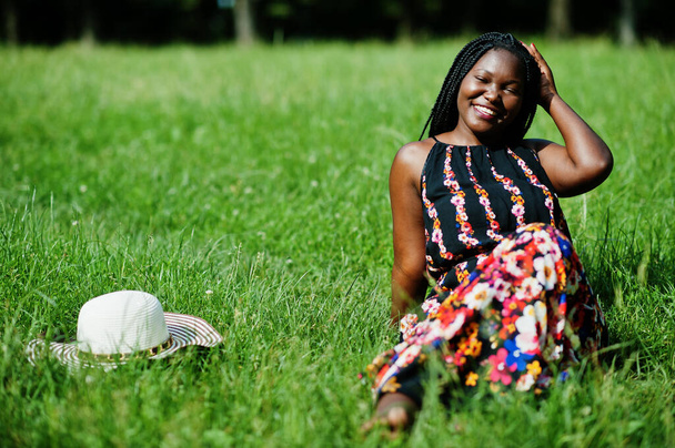 Portrait of gorgeous african american woman 20s in summer hat sitting at green grass in park. - 写真・画像