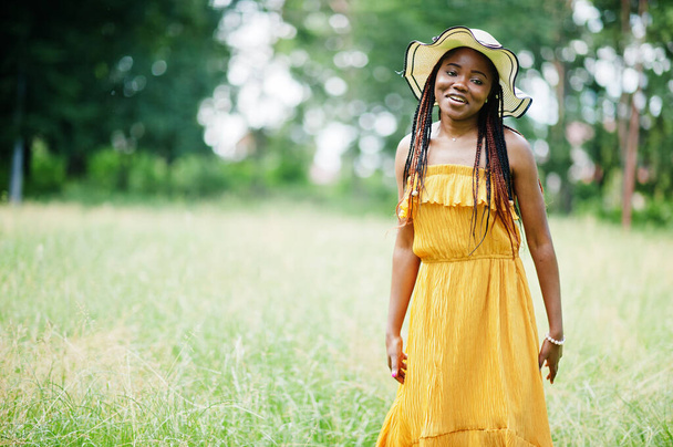 Portrait of gorgeous african american woman 20s in wear in yellow dress and summer hat posing at green grass in park. - Фото, зображення