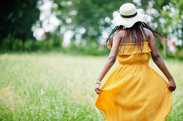 Portrait of gorgeous african american woman 20s in wear in yellow dress and summer hat posing at green grass in park. - Foto, Bild