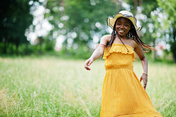 Portrait of gorgeous african american woman 20s in wear in yellow dress and summer hat posing at green grass in park. - Zdjęcie, obraz