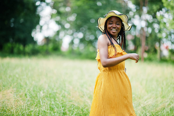Portrait of gorgeous african american woman 20s in wear in yellow dress and summer hat posing at green grass in park. - Photo, Image