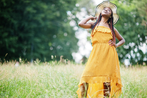 Portrait of gorgeous african american woman 20s in wear in yellow dress and summer hat posing at green grass in park. - Foto, imagen