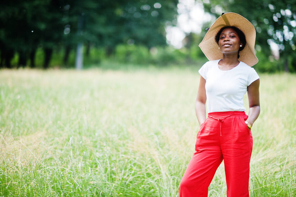 Retrato de la hermosa mujer afroamericana de 20 años usar en sombrero de verano, pantalones rojos y camiseta blanca, posando en la hierba verde en el parque. - Foto, Imagen