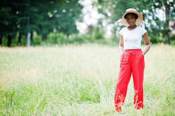 Portrait of gorgeous african american woman 20s wear in summer hat, red pants and white t-shirt, posing at green grass in park. - Foto, Bild