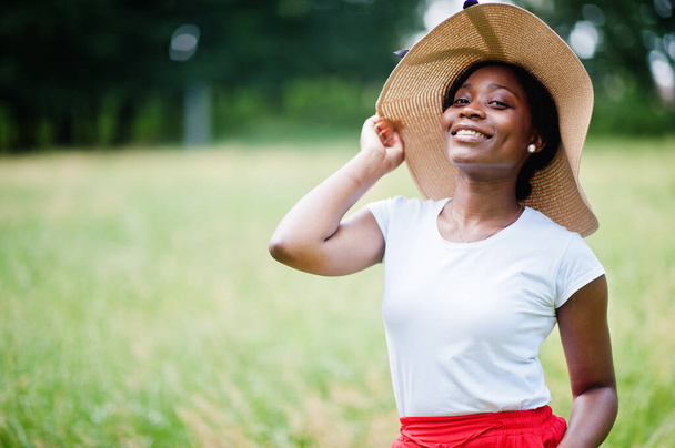 Portrait of gorgeous african american woman 20s wear in summer hat, red pants and white t-shirt, posing at green grass in park. - Фото, изображение