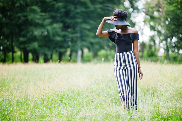 Portrait of gorgeous african american woman 20s in wear in black and white stripe pants and summer hat posing at green grass in park. - Foto, immagini