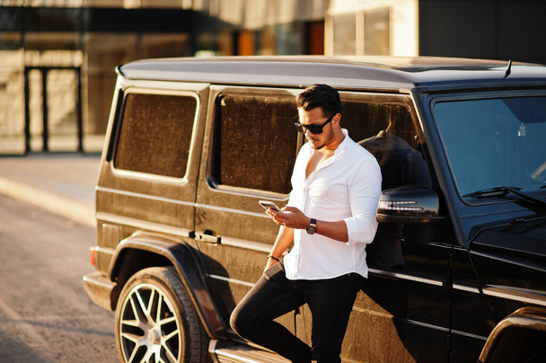 Solid asian man in white shirt and sunglasses posed near black mafia suv car with mobile phone. - Photo, image