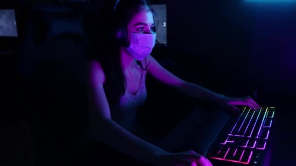 Mujer joven con auriculares y una máscara facial jugando un juego en línea en el club de juegos - Imágenes, Vídeo