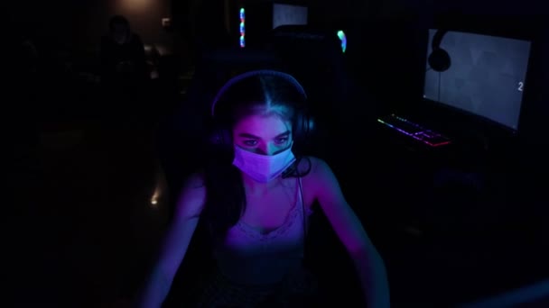 ヘッドフォンと白の顔のマスクの若い女性は、ゲームクラブでオンラインゲームをプレイ - 映像、動画