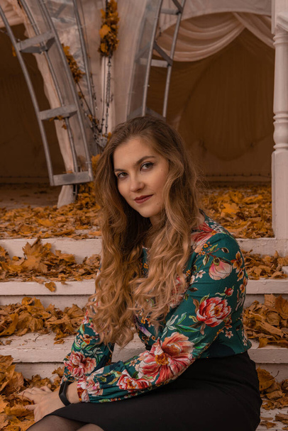 A beautiful girl stands leaning against a white railing, an adult with beautiful eyes blonde, in a colored shirt, works on a computer on the background of steps intellect autumn college standing - Foto, Imagem