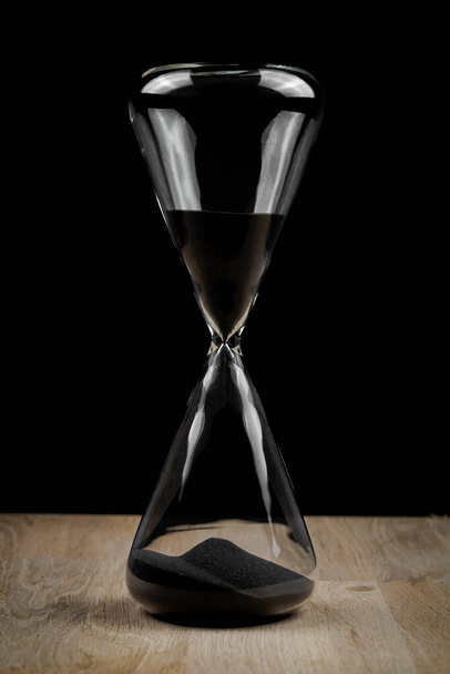 All glass hourglass against a black background on wooden surface in dramatic dark lighting. Countdown timing concept simple. - Foto, immagini