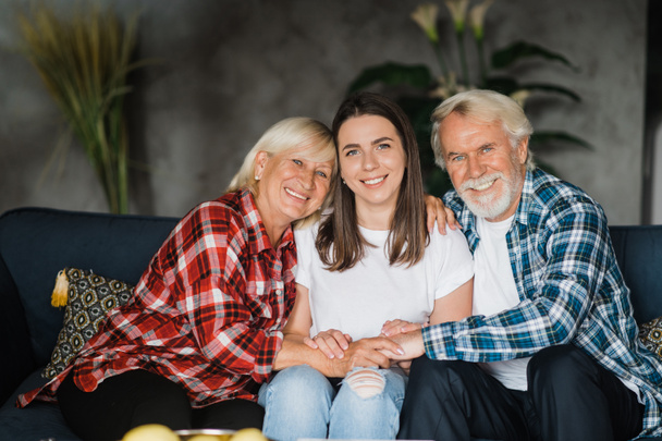 Portret rodzinny białej rodziny. Ładna córka siedzi na kanapie pomiędzy starszymi ukochanymi rodzicami, patrzą w kamerę i uśmiechają się - Zdjęcie, obraz