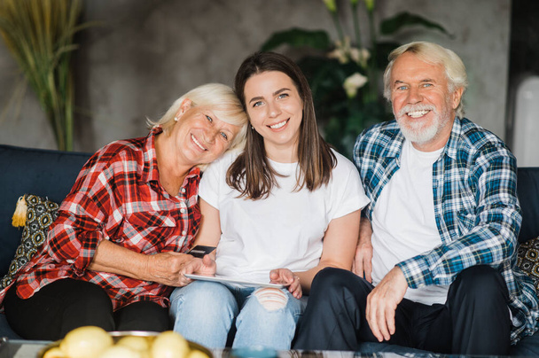 Familie portret van een blanke familie. Mooie dochter zit op de bank tussen haar bejaarde geliefde ouders, ze kijken naar de camera en lachen - Foto, afbeelding