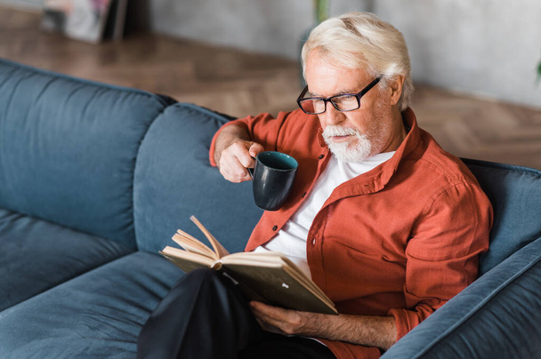 Hombre mayor de pelo gris con anteojos sentado en casa en el sofá leyendo un libro y sosteniendo una taza de té o café en la mano - Foto, Imagen