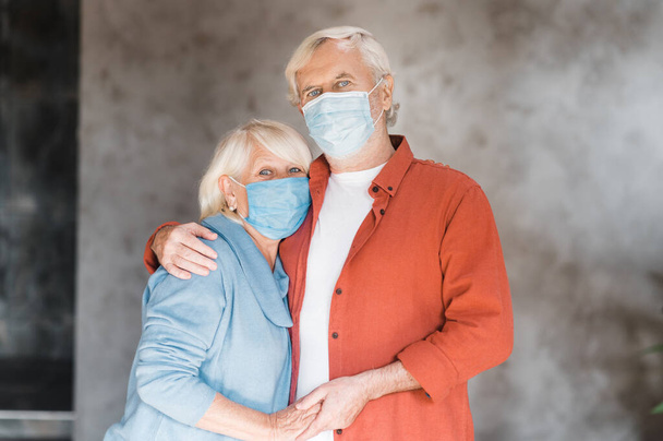 A gray-haired elderly couple in protective medical masks are hugging and looking at the camera. Pandemic - Foto, afbeelding