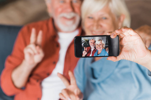 Modern öregek. Kaukázusi idős pár szelfit készít mobilon, miközben a kanapén ülnek és mosolyognak. - Fotó, kép