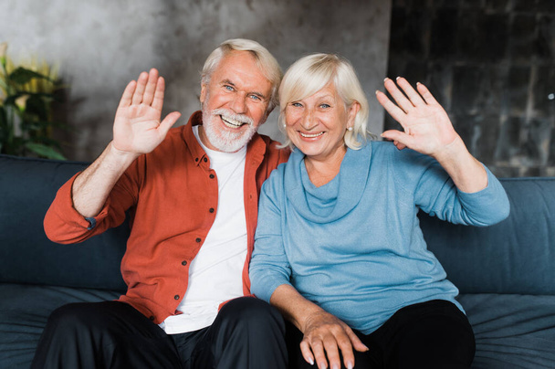Χαρούμενο χαμογελαστό καυκάσιο ζευγάρι ηλικιωμένων που κάθεται στο σπίτι κοιτάζοντας την κάμερα και κουνώντας τα χέρια κατά τη διάρκεια της επικοινωνίας με την οικογένεια ή τους φίλους με βιντεοκλήση - Φωτογραφία, εικόνα