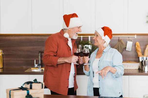 クリスマスの帽子の幸せな高齢者のカップルはリラックスしていて、ガラスのワインとキッチンで楽しんでいます - 写真・画像