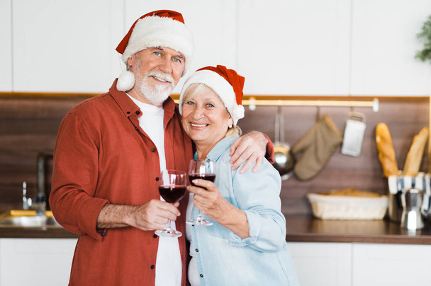 Portrait of elderly gray-haired couple in love standing in the kitchen in Christmas hats and with a glass of wine in their hands and looking at the camera with a smile - Foto, imagen