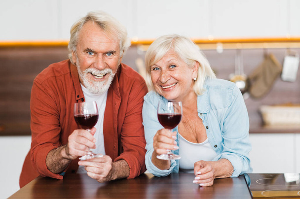 Birlikte mutluyuz. Kafkasyalı yaşlı çift ellerinde şarapla mutfakta duruyor ve gülümseyen kameraya bakıyorlar. - Fotoğraf, Görsel