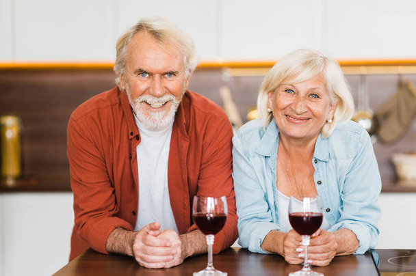 Ευτυχισμένο Καυκάσιο ζευγάρι ηλικιωμένων στέκονται στην κουζίνα με κρασί στα χέρια τους και κοιτάζουν την κάμερα χαμογελώντας - Φωτογραφία, εικόνα