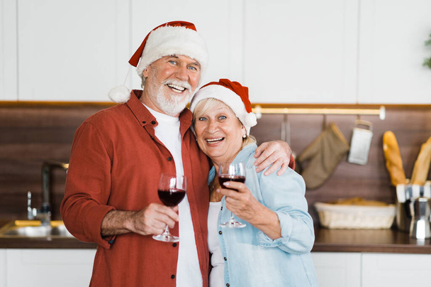 Cheerful elderly gray-haired couple in love standing in the kitchen in Christmas hats and with a glass of wine in their hands and looking at the camera with a smile - Foto, immagini