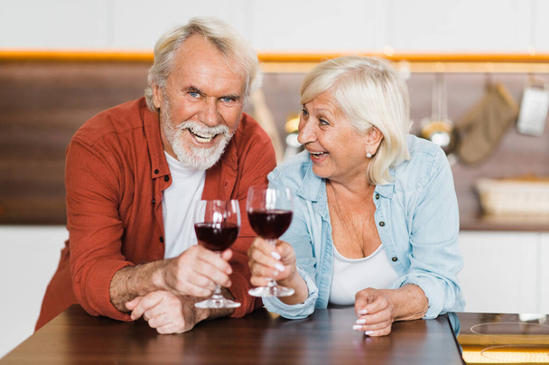 feliz canoso ancianos cónyuges en casual desgaste stand en la cocina con vino en sus manos y mirar a la cámara y riendo - Foto, imagen