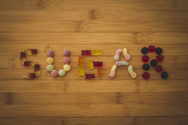 Слово сахар написано на деревянной доске с различными видами красочных кусочков конфет. Конфеты помещаются на деревянную доску с сахаром, посыпанным вокруг них. - Фото, изображение
