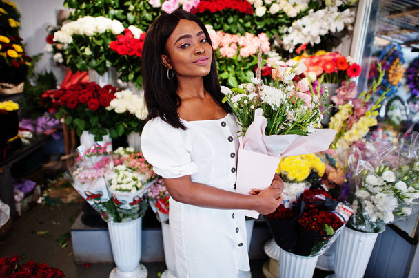 Beautiful african american girl in tender white dress with bouquet flowers in hands standing against floral background in flower shop.Black female florist. - Foto, Imagem