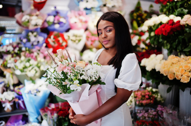 Beautiful african american girl in tender white dress with bouquet flowers in hands standing against floral background in flower shop.Black female florist. - Foto, imagen