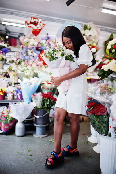 Hermosa chica afroamericana en tierno vestido blanco con flores de ramo en las manos de pie sobre fondo floral en floristería.. - Foto, Imagen