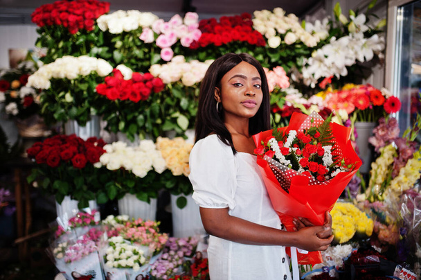 Hermosa chica afroamericana en tierno vestido blanco con flores de ramo en las manos de pie sobre fondo floral en floristería.. - Foto, imagen