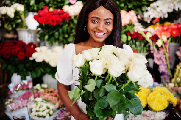 Beautiful african american girl in tender white dress with bouquet flowers in hands standing against floral background in flower shop.Black female florist. - Foto, Bild