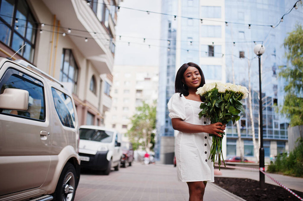 Όμορφη Αφροαμερικανή κοπέλα που κρατά μπουκέτο με λευκά τριαντάφυλλα λουλούδια κατά τη χρονολόγηση στην πόλη. Μαύρη επιχειρηματίας με πολλά λουλούδια κοντά στο αυτοκίνητο. - Φωτογραφία, εικόνα