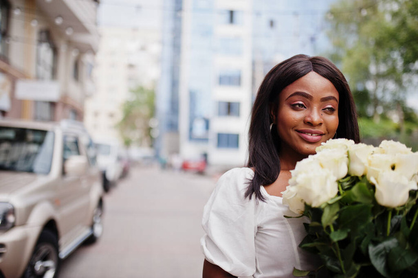 Beautiful african american girl holding bouquet of white roses flowers on dating in the city. Black businesswoman with bunch of flowers near car. - Foto, Imagem