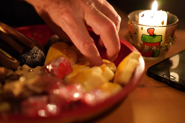 Χέρι αρπάζοντας τα Χριστούγεννα ζαχαροπλαστικής κοντά σε μαύρο κινητό τηλέφωνο σε ένα ξύλινο τραπέζι κοντά στο φως των κεριών την παραμονή των Χριστουγέννων. - Φωτογραφία, εικόνα