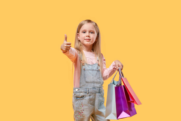 A child girl holds shopping bags in her hands and shows a thumbs-up gesture. - 写真・画像