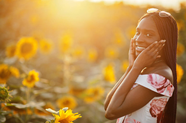 Hübsche junge schwarze Frau im Sommerkleid posiert in einem Sonnenblumenfeld. - Foto, Bild