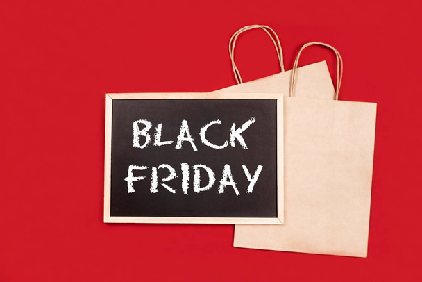 Shopping bags, chalkboard, blackboard with black friday text flat lay on red background. Black friday and online shopping concept. Big sale, low price. Advertising, marketing. Promotion design - Photo, Image