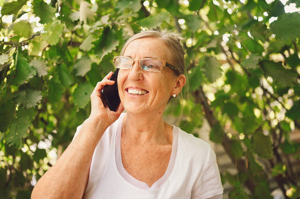 Technology, old age people concept - elderly senior old happy smiling woman in prescription eyeglasses speaks smartphone outdoor in the garden.  - Zdjęcie, obraz