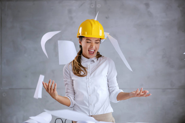 Usmívající se vzrušená architektka s helmou na hlavě dokončila svou práci včas. Všude kolem ní jsou papíry a plány.. - Fotografie, Obrázek
