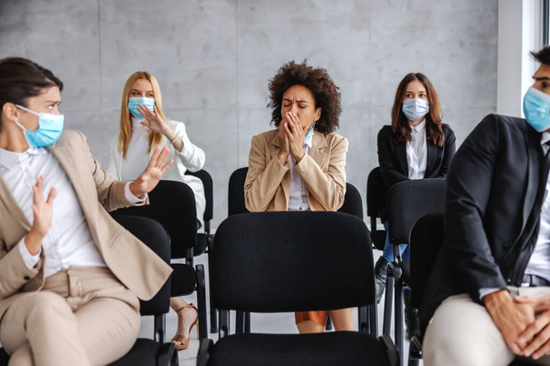 Mujer de negocios enferma tosiendo mientras está sentada con sus colegas en el seminario. Colegas temen al virus corona por lo que se esconden. - Foto, imagen