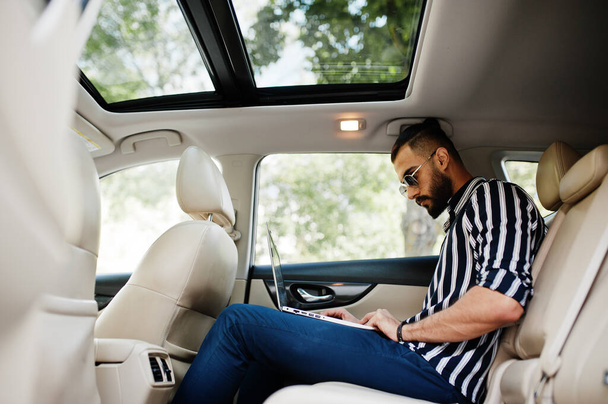 Successful arab man wear in striped shirt and sunglasses pose inside white suv car with laptop in hands. - Photo, image