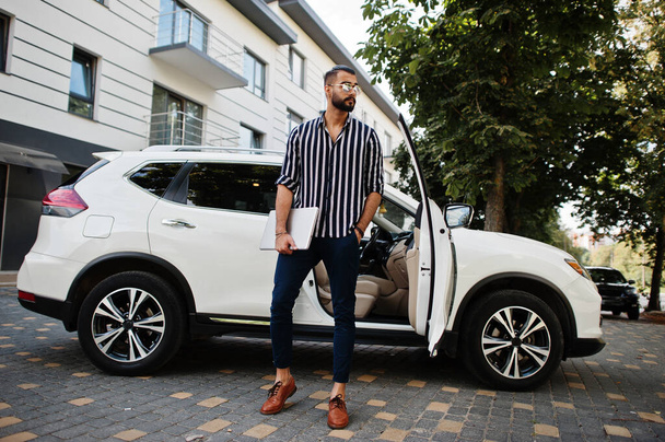 Successful arab man wear in striped shirt and sunglasses pose near his white suv car with laptop in hands. - Φωτογραφία, εικόνα