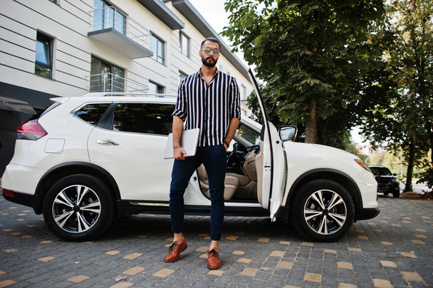 Successful arab man wear in striped shirt and sunglasses pose near his white suv car with laptop in hands. - Fotoğraf, Görsel
