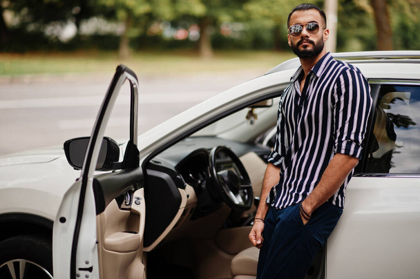 Úspěšný arabský muž nosí v pruhované košili a slunečních brýlích pózovat v blízkosti svého bílého SUV auta. Styloví arabští muži v dopravě. - Fotografie, Obrázek