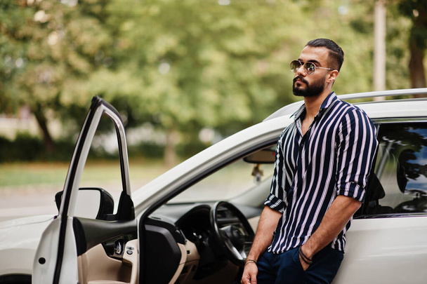 Успешная одежда араба в полосатой рубашке и солнцезащитных очках позирует возле его белого внедорожника. Транспорт в стиле арабских мужчин. - Фото, изображение